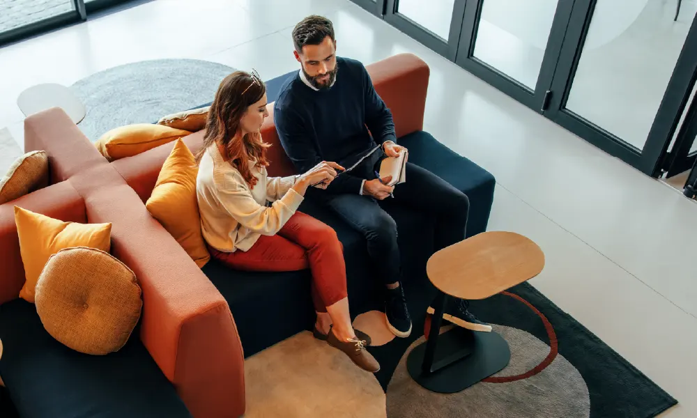 Two colleagues sit on a coral and navy couch, viewing information on a tablet